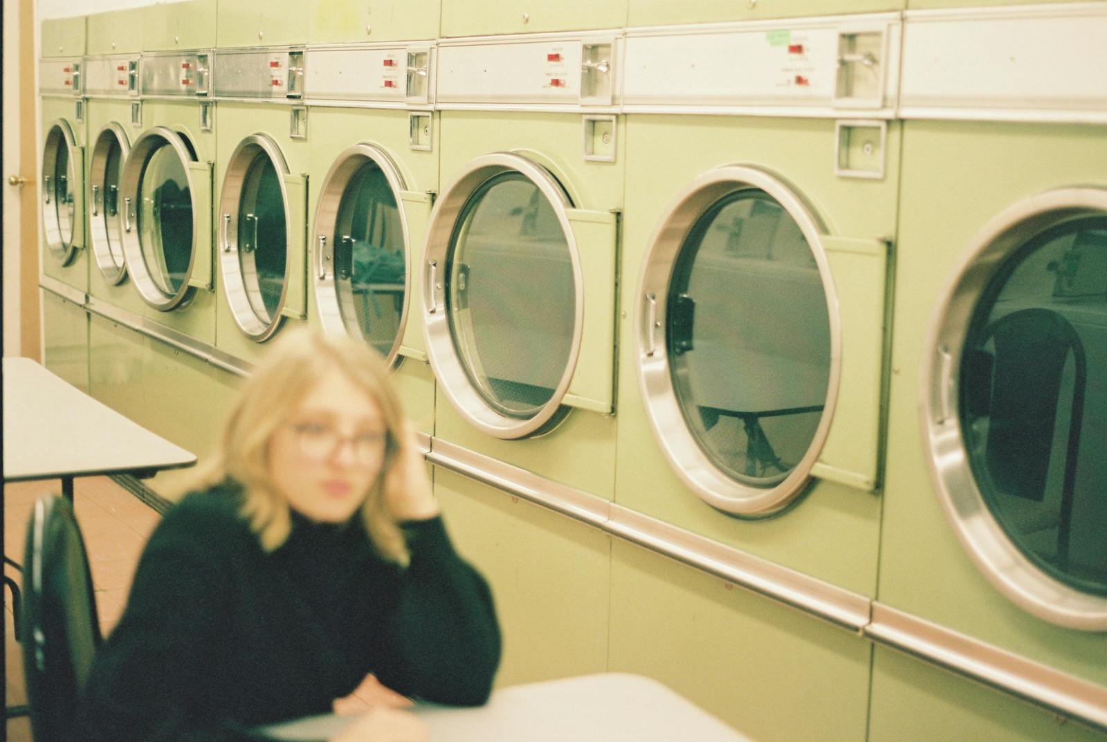 Perspective view of modern washing machines in laundry room with blurred unrecognizable female in eyeglasses sitting at table in black clothes