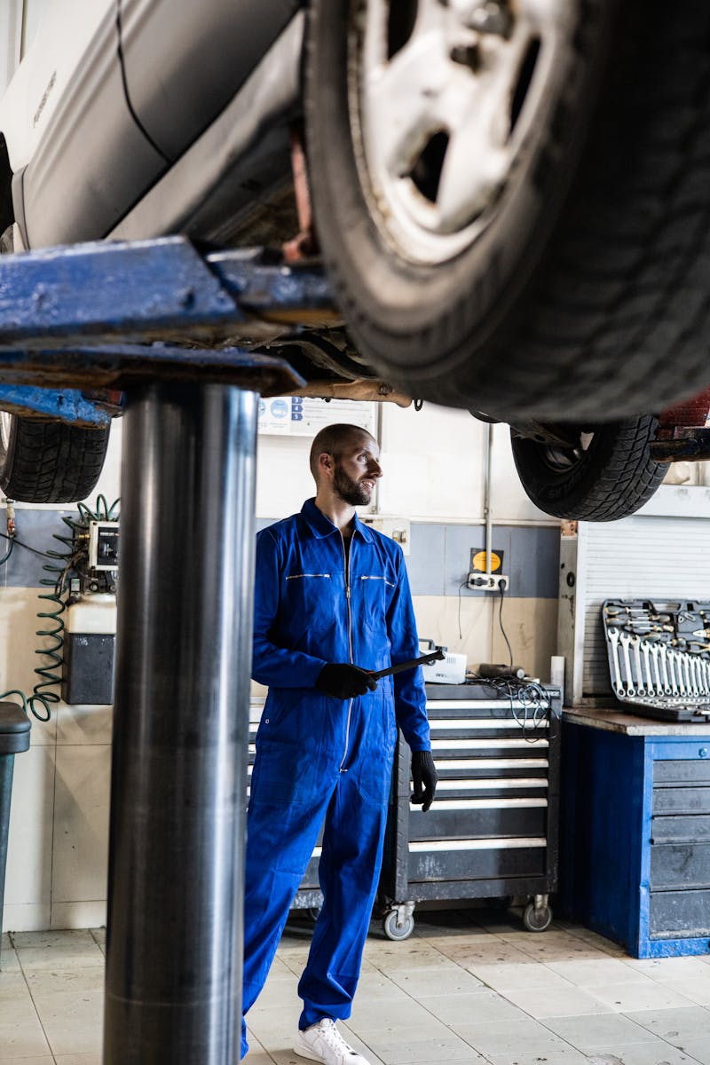 Man In Blue Coverall Standing Under A Vehicle With A Tool